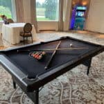 A Custom Wrapped Claw Machine sits in a room with patterned carpet, tables, chairs, large windows, and arcade games—perfect for party rentals Long Island or any fun gathering in NY.