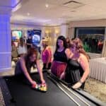 Three women stand around a pool table as one racks balls. In the background, near a Custom Wrapped Claw Machine, people enjoy the brightly lit, carpeted room—ideal for party rentals and Long Island, NY-style fun.