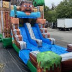 The MINECRAFT COMBO Bounce House, featuring pixelated blocks, a character face, and two blue slides, sits on outdoor pavement. A white box truck is parked nearby for delivery of the Minecraft-themed rental.