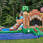 The MINECRAFT COMBO Bounce House features a slide, blocky animal heads, and pixel-style graphics, set up outdoors on grass with a blue tarp and trees in the background.