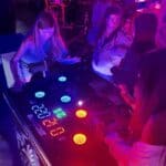 Three girls play an interactive arcade game with colorful, illuminated buttons in a dimly lit room with purple lighting. Perfect for party rentals Long Island, the setup features a digital scoreboard on the game console.