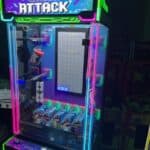 A brightly lit arcade game machine called "Brick Attack" features neon blue, green, and pink lights. The screen displays a grid, and there are prizes inside, visible through a glass panel—perfect for NY party rentals Long Island events.