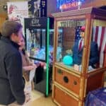 Two people smile in front of a “TRUMP TELLS ALL” fortune teller machine at an NY arcade. Nearby, a claw machine and a festive display add to the fun—perfect inspiration for party rentals Long Island style.