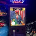 A fortune teller machine with a mannequin resembling Donald Trump inside, wearing a red "Make America Great Again" hat, suit, and tie—a fun attraction for NY party rentals Long Island. The sign above reads "TRUMP TELLS ALL" in bright neon lights.