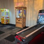 A row of arcade machines is set against a curtain, including a claw machine, a Zoltar fortune teller, and a Skeeball game—perfect for party rentals Long Island— all illuminated by soft indoor lighting.