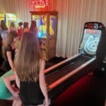 Two girls stand at the start of a Skee-Ball lane in an arcade. Behind them, a group gathers near a Zoltar fortune teller machine and other bright arcade games, perfect for party rentals Long Island events, all set against a white curtain backdrop.