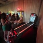 A group of young people play skee-ball in a dimly lit arcade, perfect for party rentals Long Island. One person rolls a ball toward the machine as others watch. Behind them are more games, including a Zoltar fortune teller.