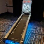 A skee-ball arcade machine in NY is illuminated with blue lights, ready to play. The lane leads up to numbered target rings, with balls in front and a patterned carpet below—perfect for party rentals Long Island fun.