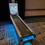 A skee-ball arcade game machine is placed on a carpeted floor with blue accent lighting, perfect for NY party rentals Long Island events, featuring a ramp and target rings marked with different point values.