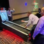Three men in dress shirts and slacks play a skee-ball arcade game in a lively room with colorful carpet and decor. It’s the perfect scene for party rentals Long Island, NY, where friends gather to cheer and enjoy arcade fun together.