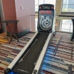 A Skee-Ball arcade game machine sits on a colorful carpet in a spacious, sunlit room—perfect for party rentals Long Island, NY—with blue balls stacked at the end and a small round table nearby.