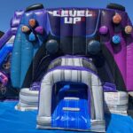 A large, colorful inflatable slide shaped like a video game controller, with "LEVEL UP" written at the top, sits outdoors on a blue tarp under a sunny sky—perfect for party rentals Long Island, NY.