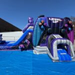 A large inflatable obstacle course and slide with a video game theme, featuring "LEVEL UP" and "GAME OVER," set up outdoors on a sunny day with a blue tarp—perfect for bounce house Long Island or party rentals Long Island, NY.