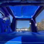 View from inside a blue bounce house Long Island with mesh windows, showing climbing holds on the left wall and a clear sky with trees outside—perfect for party rentals Long Island, NY.