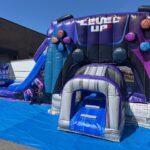 An inflatable bounce house Long Island shaped like a large video game controller in purple, blue, and black, labeled "LEVEL UP," set up outdoors on a blue tarp. A slide and entrance tunnel are visible—perfect for NY party rentals!.