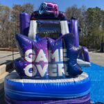 An inflatable obstacle course with purple and blue colors features "GAME OVER" in large white letters and a video game controller graphic at the top. Perfect for NY events, this party rentals Long Island favorite is set up outdoors near trees and pavement.