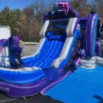 A large inflatable water slide with a blue and purple gaming theme, featuring "GAME OVER" on the side, is set up outdoors on a sunny day. Perfect for party rentals Long Island, it has multiple lanes and sits on blue and black tarps.