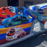 Colorful inflatable obstacle course themed around race cars, featuring a blue and green car with the number 99 and vibrant racing graphics. Perfect for party rentals Long Island, NY—this fun bounce house brings excitement to any outdoor event.
