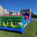 A colorful bounce house Long Island inflatable obstacle course is set up on a grassy backyard, with two entry holes in front. Suburban houses and a white fence are visible in the background under a clear blue sky.