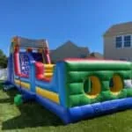 A large, colorful inflatable obstacle course and slide—perfect for party rentals Long Island, NY—stands on a grassy backyard, with two-story houses and a clear blue sky in the background.