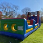 A large, colorful inflatable obstacle course from party rentals Long Island, NY sits on green grass in a sunny park, with trees and a building visible in the background.
