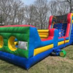 A large, colorful inflatable obstacle course with climbing walls and crawl-through holes sits on a grassy lawn, perfect for party rentals Long Island, NY, surrounded by bare trees and a clear sky.