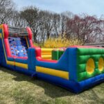 A colorful inflatable obstacle course with climbing and slide sections is set up on grass in a backyard, perfect for party rentals Long Island, NY, surrounded by trees and a nearby house.