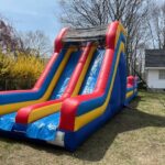 An inflatable slide with red, blue, and yellow sections is set up on a grassy yard in NY, with trees and a house in the background on a sunny day—perfect for bounce house Long Island party rentals.