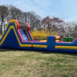 A large, colorful inflatable obstacle course from party rentals Long Island sits on a grassy lawn, surrounded by trees and bushes. The course features a climbing wall, slide, and fun barriers in bright blue, yellow, and red sections.