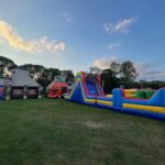 Colorful inflatable obstacle courses and games, including a pumpkin-themed bounce house Long Island, are set up on a large grassy field under a blue sky with scattered clouds, surrounded by trees and a building in NY.