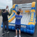 Two people play a basketball shooting game on an inflatable court outdoors, part of a bounce house Long Island setup. One in a "STAFF" shirt and both aim basketballs at hoops, perfect for party rentals Long Island, NY gatherings.