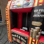 Inflatable carnival game booth with slot machine graphics, the words "Match 3 & Win" and "Go for the Jackpot," and stacks of gold coins. Perfect for party rentals Long Island, NY; a chair and table are visible inside the booth.
