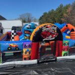 A colorful inflatable obstacle course with a racing and monster truck theme, perfect for NY party rentals Long Island, featuring images of trucks, a "Speed Way" sign, and tunnels and barriers—set outdoors near trees and buildings.