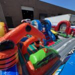 Colorful inflatable obstacle course with race car and tire designs, perfect for party rentals Long Island, NY; set up outdoors near a warehouse and white trailer on a sunny day.