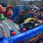 Colorful inflatable obstacle course with a race car theme, perfect for NY events—features blown-up cars, ramps, and a checkered "Finish" arch. Ideal for party rentals Long Island, set outdoors near industrial buildings on a sunny day.