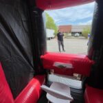 View from inside a red and black bounce house Long Island, looking out at a white folding chair and a man standing in a parking lot with trees, cars, and a building in NY under a partly cloudy sky.