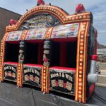 An inflatable carnival game booth themed like a slot machine, labeled "Jumbo Slot," with three game stations and bright lights—perfect for party rentals Long Island, NY—set up outdoors on a black mat near a building and metal bleachers.