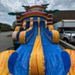 A colorful inflatable water slide with blue and yellow lanes, palm trees on top, and a small pool at the bottom is set up outdoors near a building and parked vehicles under a cloudy sky—perfect for party rentals Long Island, NY.