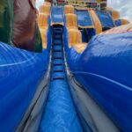 A low-angle view of a large inflatable slide with bright blue, yellow, and orange colors, and palm tree decorations at the top, under a partly cloudy sky—perfect for party rentals Long Island fun.