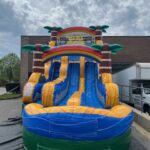 An inflatable water slide with palm tree decorations and two blue sliding lanes is set up outdoors near a building and a parked truck under a partly cloudy sky—perfect for party rentals Long Island, NY.