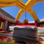 A colorful inflatable obstacle with yellow and red arches stands under a clear blue sky. Perfect for NY events, this bounce house Long Island features cushioned platforms and a central padded post, resembling a giant game arena.