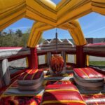 Colorful inflatable game structure with a central hanging punching bag and large padded platforms, perfect for party rentals Long Island, set outdoors on a sunny day with trees and a building in the background.