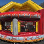 A colorful inflatable bounce house Long Island designed to resemble a pizza, with red, yellow, and gray colors, cheese and sauce details, and mesh windows; it is set up outdoors on a sunny day.