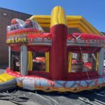 An inflatable bounce house Long Island with a volcano and lava theme sits outdoors on a tarp. The words "FLOOR IS LAVA" are visible. Perfect for party rentals Long Island, it features red, yellow, and orange to resemble flowing lava.