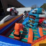 Colorful inflatable obstacle course with car-themed designs, perfect for NY events. Set up outdoors near a brick building, this bounce house Long Island rental features cars and racing elements—a fun party rentals Long Island choice!.