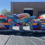 Colorful inflatable obstacle course with race car graphics, featuring large 3D cars numbered 99 and 13, plus a “The Ultimate Garage” sign—perfect for party rentals Long Island. Set up outdoors near a brown building.