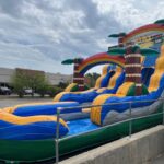 A large, colorful inflatable water slide with palm tree decorations—perfect for party rentals Long Island—is set up outdoors near a concrete ramp and metal rail, with buildings and a cloudy sky in the background.