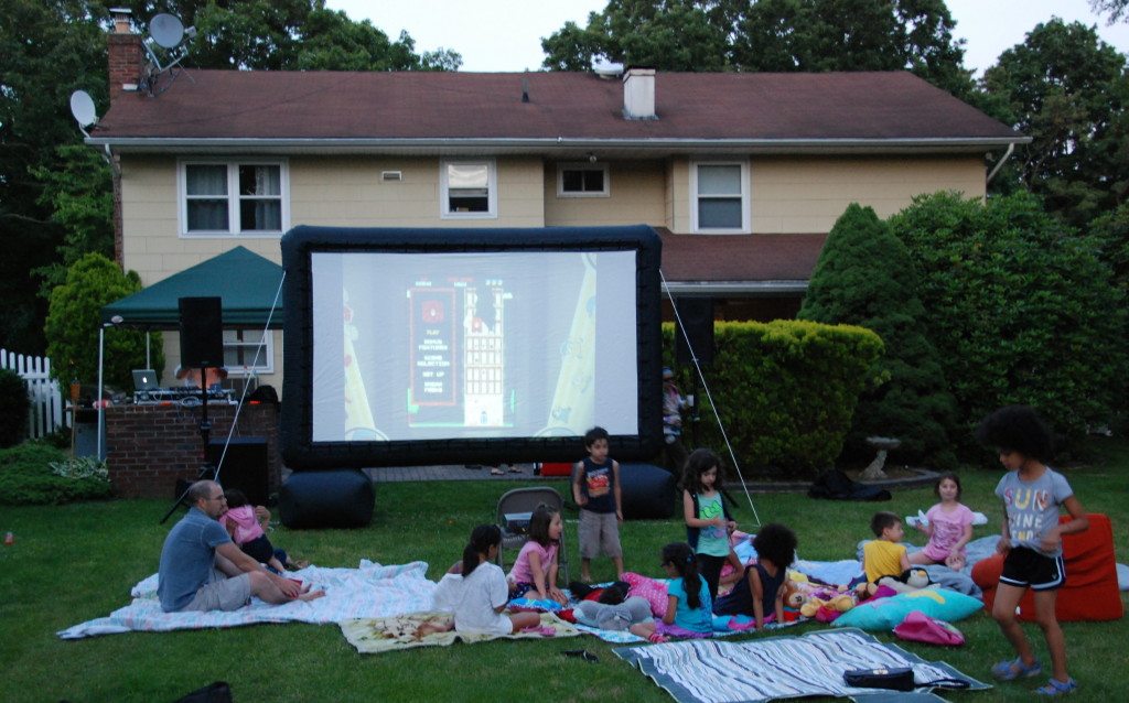 A group of children and adults sit on blankets and pillows on a lawn, watching a movie projected on an outdoor screen in front of a house during the evening. Some kids are playing and walking around.