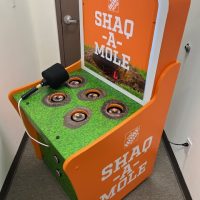 An orange arcade cabinet labeled "SHAQ-A-MOLE" resembling a whack-a-mole game, with a black foam mallet and holes featuring miniature basketball hoops, placed in a hallway near a closed door.