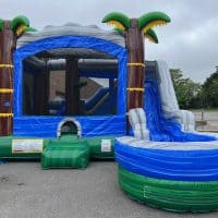 A large inflatable bounce house with a blue and gray slide, decorated with palm trees, is set up outdoors on pavement near a building and greenery.